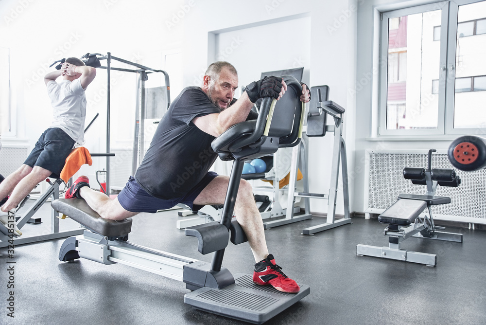Adult gray-haired man trains on fitness equipment in the gym, pumps legs and arms muscles, loses weight. Concept of healthy lifestyle in old age Adult gray-haired man trains on fitness equipment in the gym, pumps legs and arms muscles, loses weight. Concept of healthy lifestyle in old age