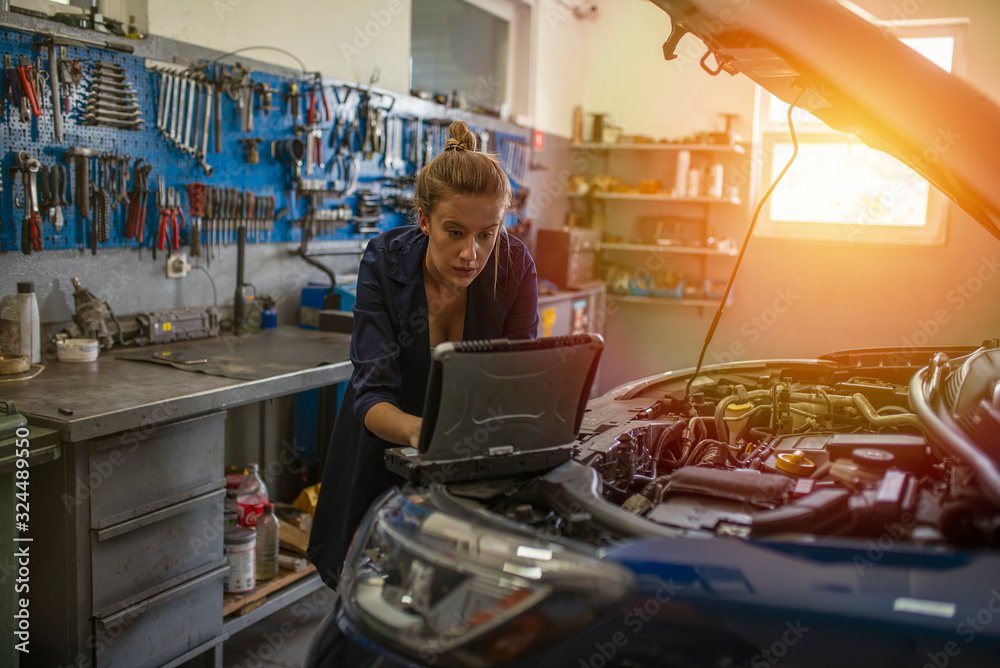 An attractive woman mechanic working on a car in a repair shop. A female mechanic is working under a bonnet of a car in a garage repair shop. She is wearing blue overalls.