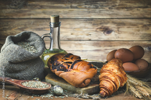 Sweet bread, croissant with poppy seeds, wheat ear, egg, and grain in a bag lie on a wooden rustic table against a wooden background. Low key. Toned. Focus concept
