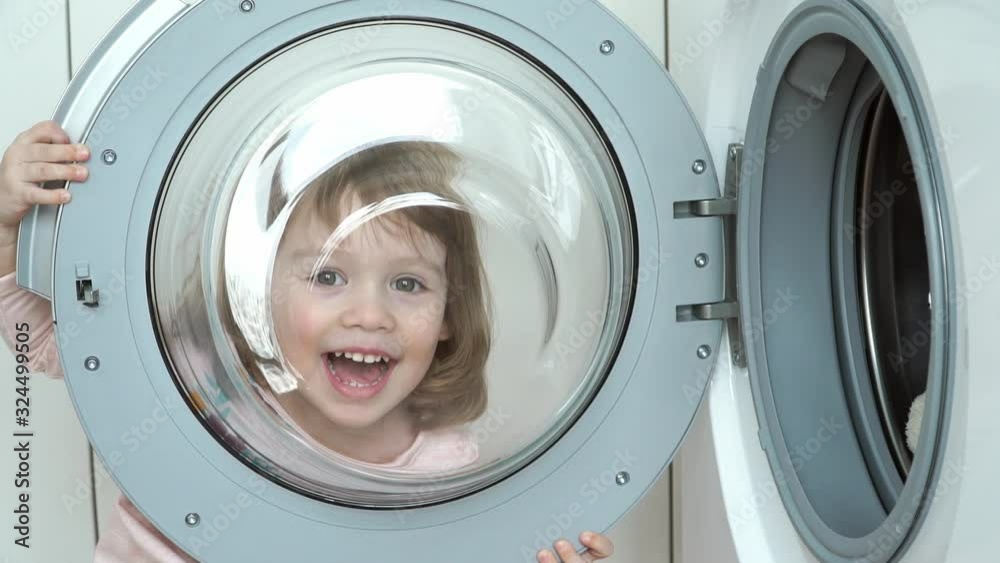 Close up of little funny 3 year old girl plays with hatch of washing machine. Child looks through hatch, fools around, smiles and laughs