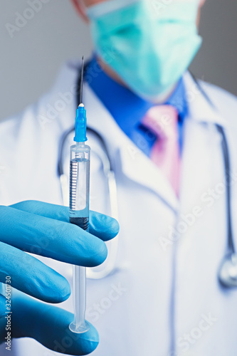 Close up view of medical syringe in rubber blue glove dressed hand, man chest in white medical robe with stethoscope on a blurred background