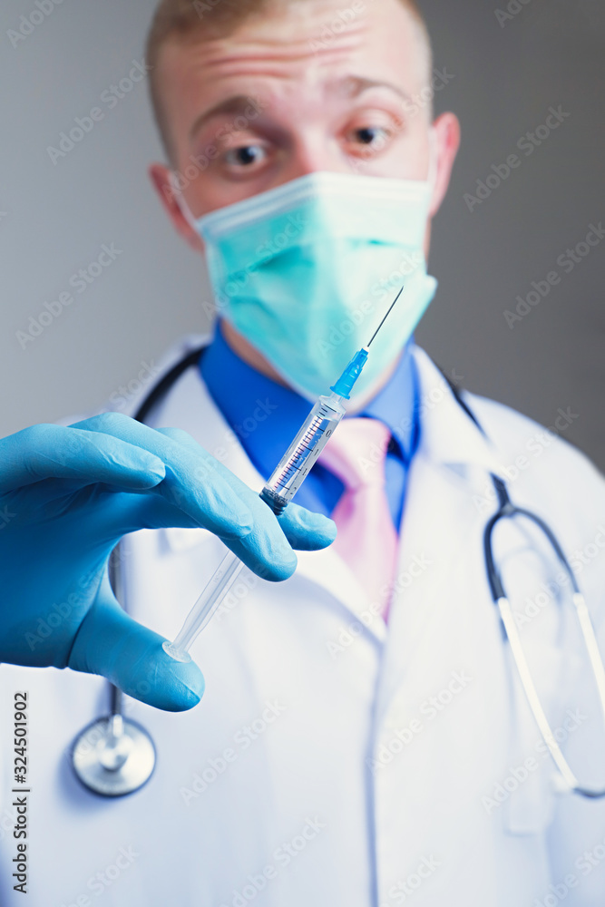 Scared young doctor in gauze mask and medical robe holding syringe in his blue rubber glove dressed hand. Intern education concept Scared young doctor in gauze mask and medical robe holding syringe in his blue rubber glove dressed hand. Intern education concept