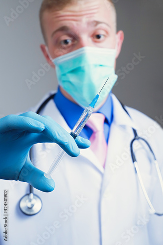Scared young doctor in gauze mask and medical robe holding syringe in his blue rubber glove dressed hand. Intern education concept