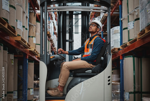 Happy forklift driver focused on carefully transporting stock from shelves around the floor of a large warehouse wearing a white helmet and vest photo