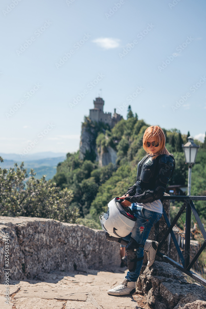 Woman posing. Girl dressed in a motorcycle outfit and sunglasses. body protection turtle and knee pads, helmet in hand. Fortress, stone steps, stairs. Vertical photo. Pass of the witches San Marino