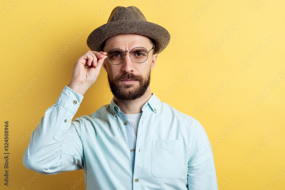 Portrait of brunette man with beard wearing in shirt, hat and glasses. He frown eyesbrows and looking at the camera touching glasses. Portrait of brunette man with beard wearing in shirt, hat and glasses. He frown eyesbrows and looking at the camera touching glasses.