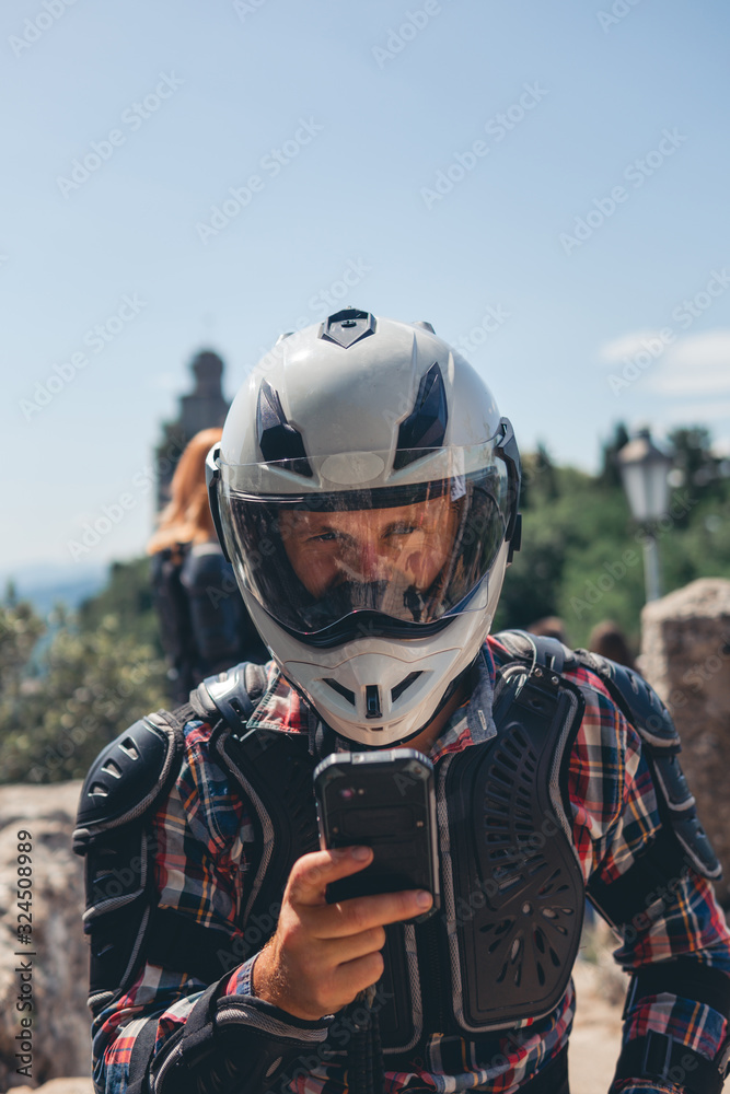 Man dressed in a motorcycle outfit and white helmet and sunglasses. body protection turtle. Fortress on background. Stylish. close up. use smartphone. San Marino, Italy Man dressed in a motorcycle outfit and white helmet and sunglasses. body protection turtle. Fortress on background. Stylish. close up. use smartphone. San Marino, Italy