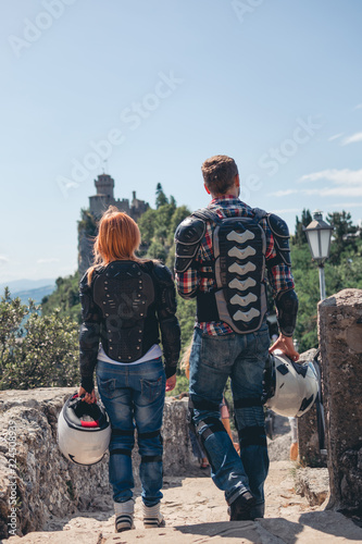 Back view motorcyclists couple. Dressed in a motorbike outfit and sunglasses. body protection turtle and knee pads, holding helmets in hand. Fortress on background. Vertical photo. San Marino