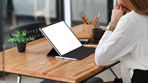 Photo of young beautiful woman working as secretary sitting and concentrated on white blank screen computer tablet with keyboard case at the modern wooden table with orderly office as background.