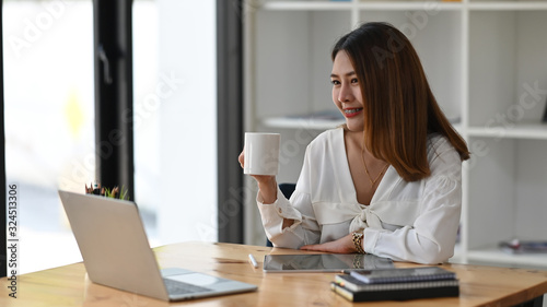 Beautiful woman working as secretary holding a coffee cup in hand while sitting in front the computer laptop at the modern working table with orderly office as background.