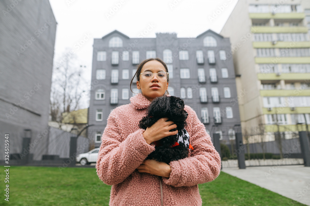 Portrait of stylish lady in glasses standing on the street with a curly little dog and posing on camera wearing a pink coat. Attractive girl walking with toy poodle in her hands on city streets. Pets