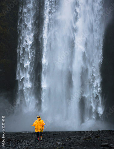 Tourist wearing a yellow raincoat walks to the Skogafoss waterfall in Iceland