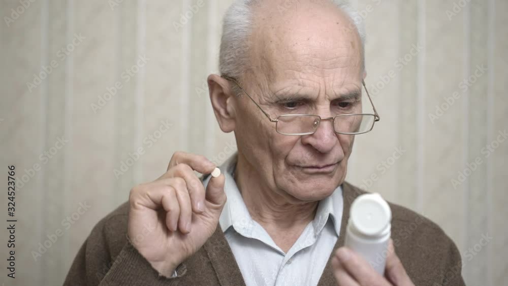 serious senior man with glasses looks at white plastic bottle of pills reading instruction manual against room wallpaper serious senior man with glasses looks at white plastic bottle of pills reading instruction manual against room wallpaper