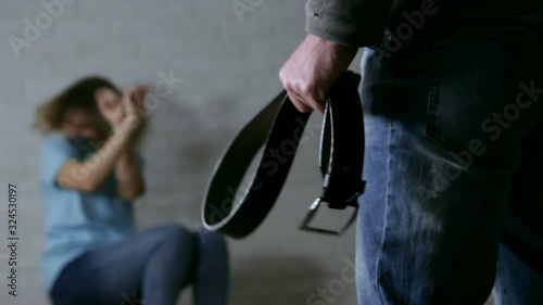 Woman trying to hide herself while man standing with belt in hand - domestic violence, abuse and people concept