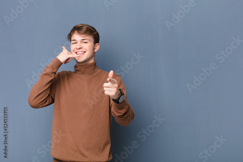 young blonde man smiling cheerfully and pointing to camera while making a call you later gesture, talking on phone isolated against flat wall photo