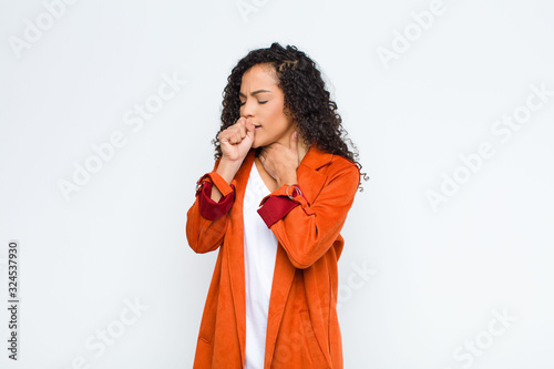 young black woman feeling ill with a sore throat and flu symptoms, coughing with mouth covered against white wall