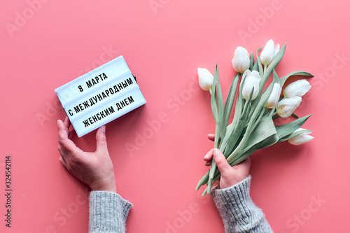 Flat lay, woman hands with lightboard and white tulip flowers on vibrant pink background. Text in Russian on ligtht board means March 8 international women day. Springtime festive greeting. photo