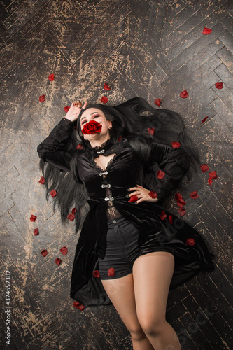 A happy plus size model in a black dress with long hair lies on a wooden floor. Young plump woman posing on camera in rose petals. The body is positive. The concept of one beauty. XXXL fashion.