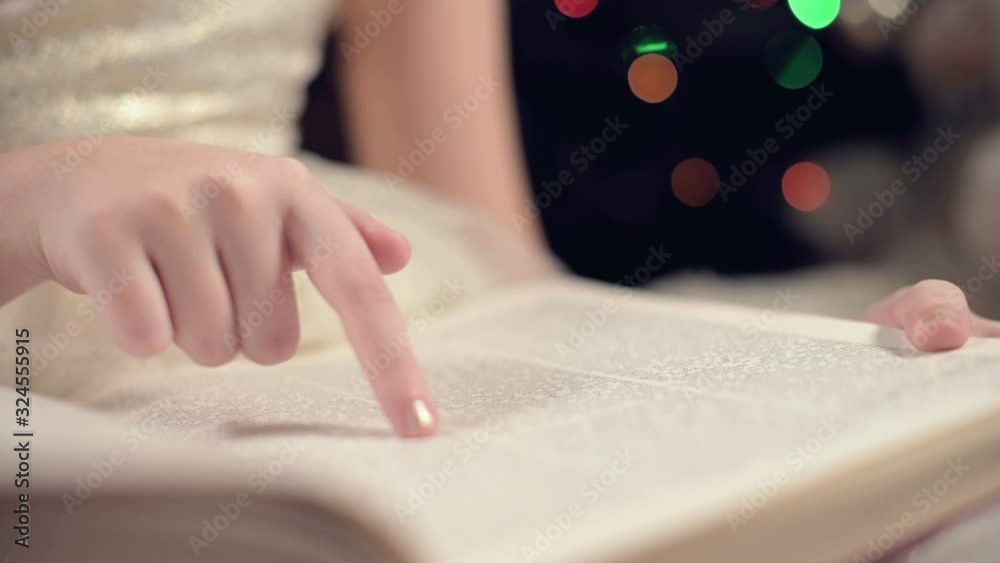 Close-up A little blonde girl in a festive dress with a book in her hands sits next to soft toys against the background of a Christmas tree and reads a book leading the page with her place. Close-up A little blonde girl in a festive dress with a book in her hands sits next to soft toys against the background of a Christmas tree and reads a book leading the page with her place.
