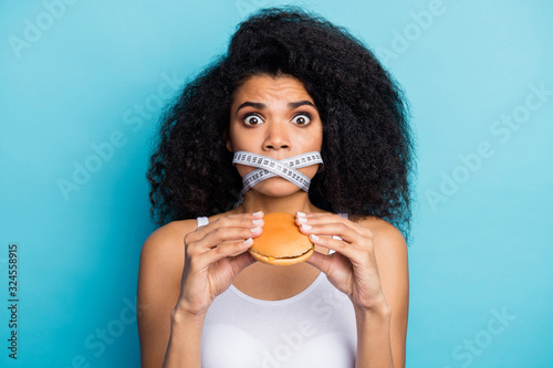 Close-up portrait of nice attractive scared wavy-haired girl holding in hand burger restriction mouth crossed closed by centimeter line isolated on bright vivid shine vibrant blue color background