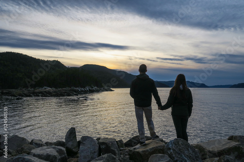 Back view of a young couple standing on rocks while holding hands and looking at the sunset, in Petit-Saguenay, Quebec, Canada © jonas