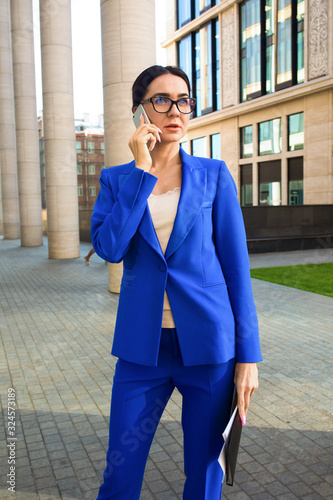 Woman in glasses and formal suit confident real estate agent talking with client via mobile phone and holding in hand contract while standing outdoors near modern penthouse building