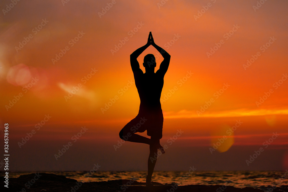 Fit young man practices vrikshasana yoga on the summer beach at sunset.