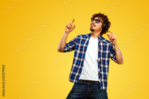 Handsome happy european man with beard in blue shirt smiling and dancing isolated on yellow background. Guy in headphones listening to music. Lifestyle concept.