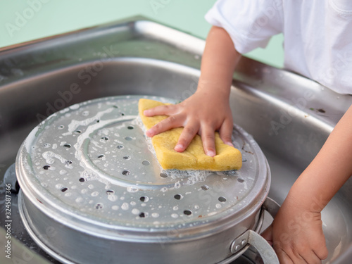 Little child hand washing pot with yellow sponge at sink in kitchen. Kid learning to clean pot, doing housework. ef concept.
