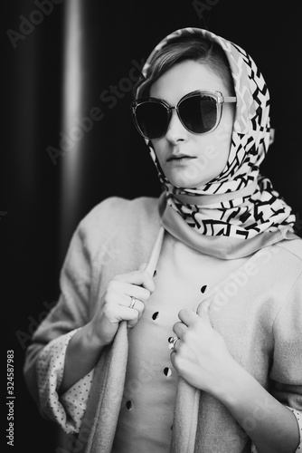 Elegant fashion. Stylish blonde woman in pink clothes, scarf and sunglasses posing in the city against a background of black huge barrels. Soft selective focus. Black-white portrait.