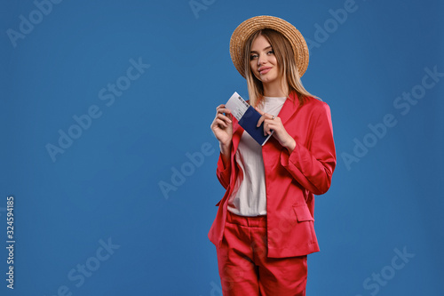 Blonde lady in straw hat, white blouse, red pantsuit. She is holding passport and ticket, posing on blue background. Travelling concept. Close-up © nazarovsergey