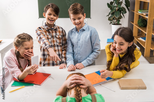 selective focus of schoolkid with smartphone and cruel classmates laughing near bullied kid, cyberbullying concept photo