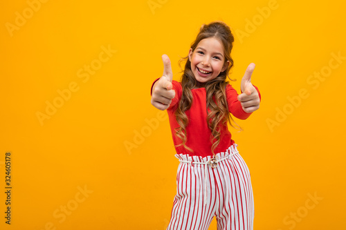 charming teen girl in a red blouse and striped trousers shows a super class on a yellow background with copy space