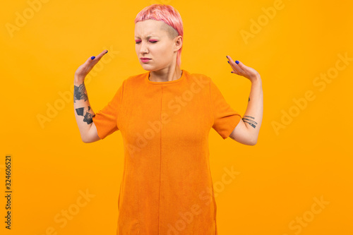 Extraordinary beautiful woman with short pink hair and big tattoo on her hand doesn t understand something isolated on orange background