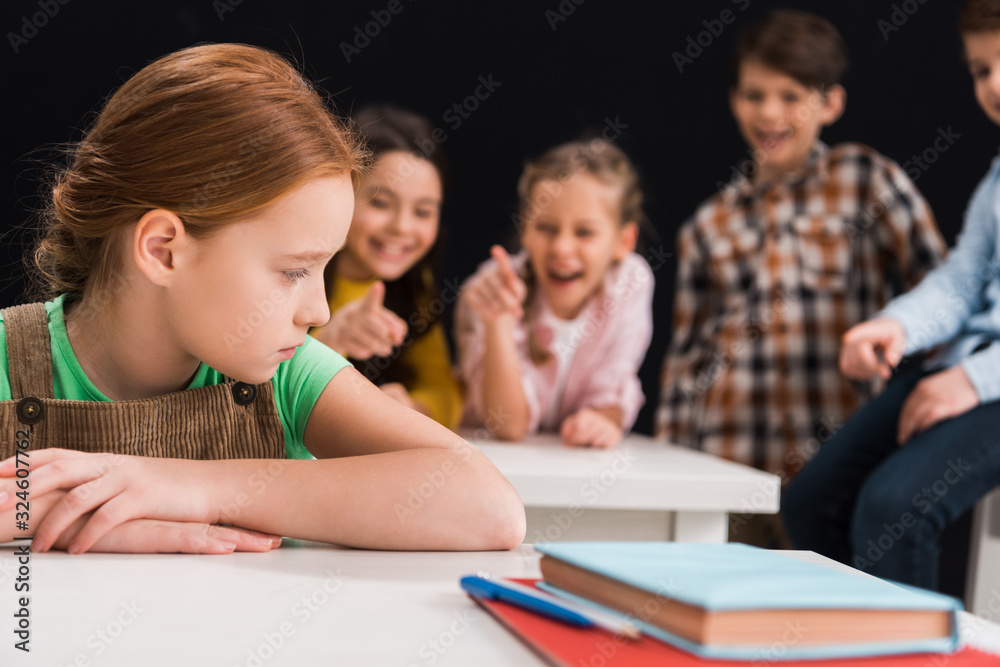 selective focus of upset schoolkid sitting near classmates laughing and pointing with fingers isolated on black, bullying concept selective focus of upset schoolkid sitting near classmates laughing and pointing with fingers isolated on black, bullying concept