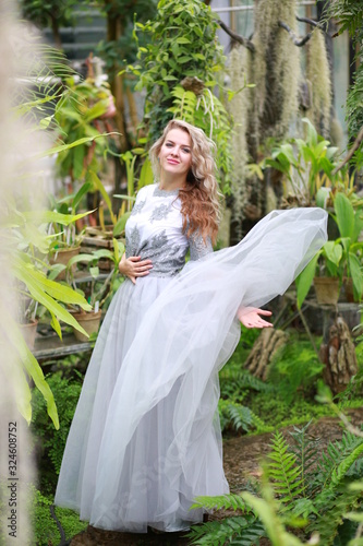  beautiful blonde long hair girl in a flying tulle skirt walks in a botanical garden among green plants