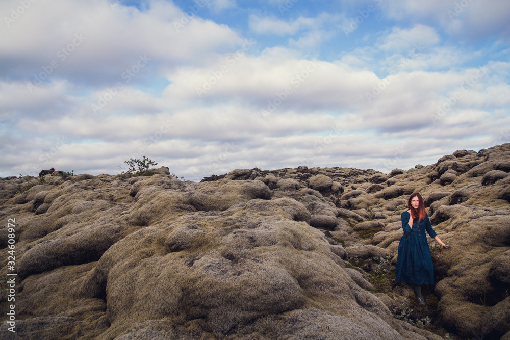 Young beautiful woman in blue dress stands on the volcanic formations covered by green moss on a background sky. Place for text or advertising