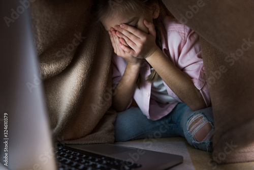 selective focus of frustrated kid covered in blanket covering face while crying near laptop, cyberbullying concept