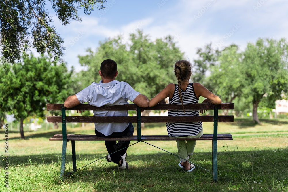 Couple sitting on a wooden bench under a tree looking forward Couple sitting on a wooden bench under a tree looking forward