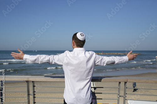 young Jew in kippah hat stands with outstretched arms on a background of blue sea and blue sky