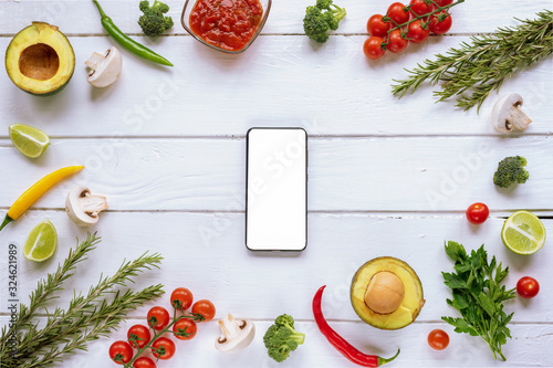 Ingredients for Italian food: tomatoes, mushrooms, broccoli, chilli peppers, avocado, rosemary and parsley on a white wooden background.  Mobile phone with blank screen Top view, flat lay, copy space. photo