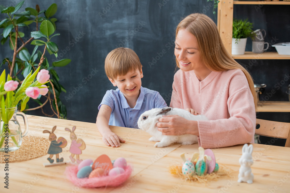Fototapeta premium Smiling mother stroking fluffy rabbit together with excited son while celebrating Easter