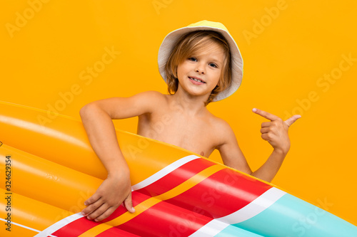 Handsome boy in swimming trunks holds a rubber mattress, smiles and gesticulates isolated on orange background photo