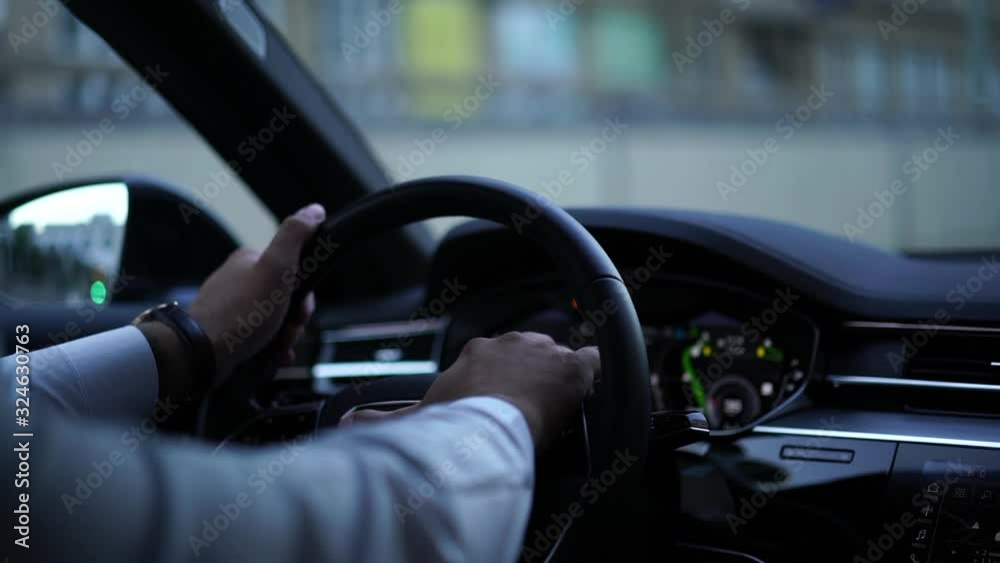 Cropped view from back seat in slow motion of business man holding hands on wheel driving modern automobile on urban city road follow rules and safety during transportation. Confidence in steering Cropped view from back seat in slow motion of business man holding hands on wheel driving modern automobile on urban city road follow rules and safety during transportation. Confidence in steering
