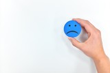Hand holding a blue sad face icon in white background with copy space. Sadness, bad customer service and unsatisfied client.
