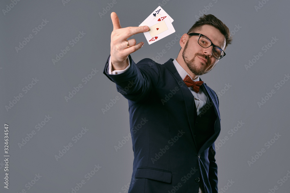 Man in black classic suit, red bow-tie, glases is showing two playing cards, posing on gray studio background. Gambling, poker, casino. Close-up. Man in black classic suit, red bow-tie, glases is showing two playing cards, posing on gray studio background. Gambling, poker, casino. Close-up.