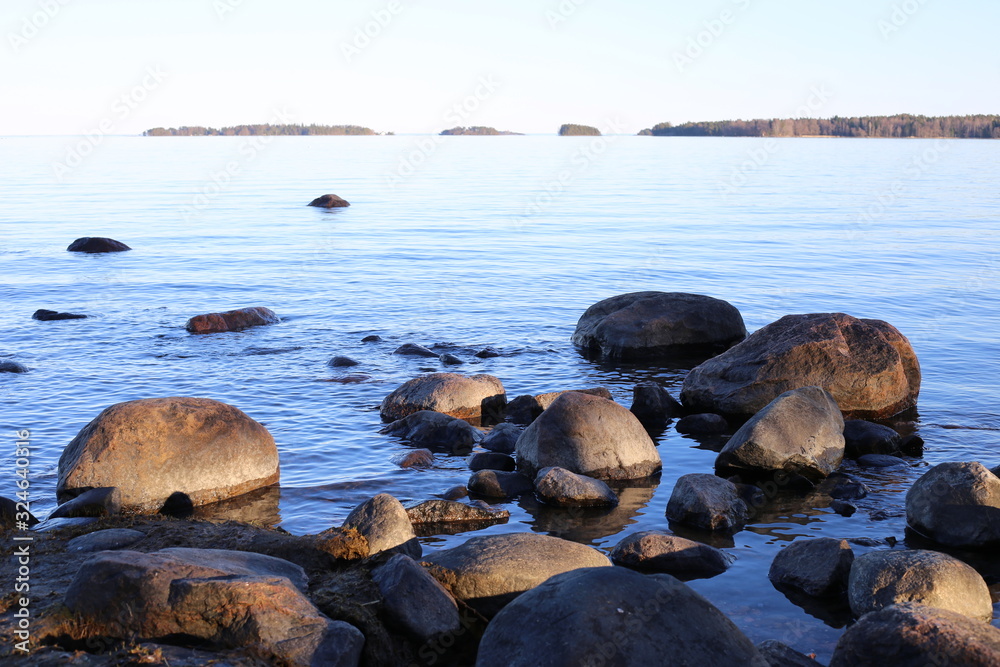 Beautiful calm Baltic Sea photographed in Espoo, Finland during a sunny summer day. The beach has a lot of medium sized rocks. You can also see some islands with forest in the horizon. Clear sky.