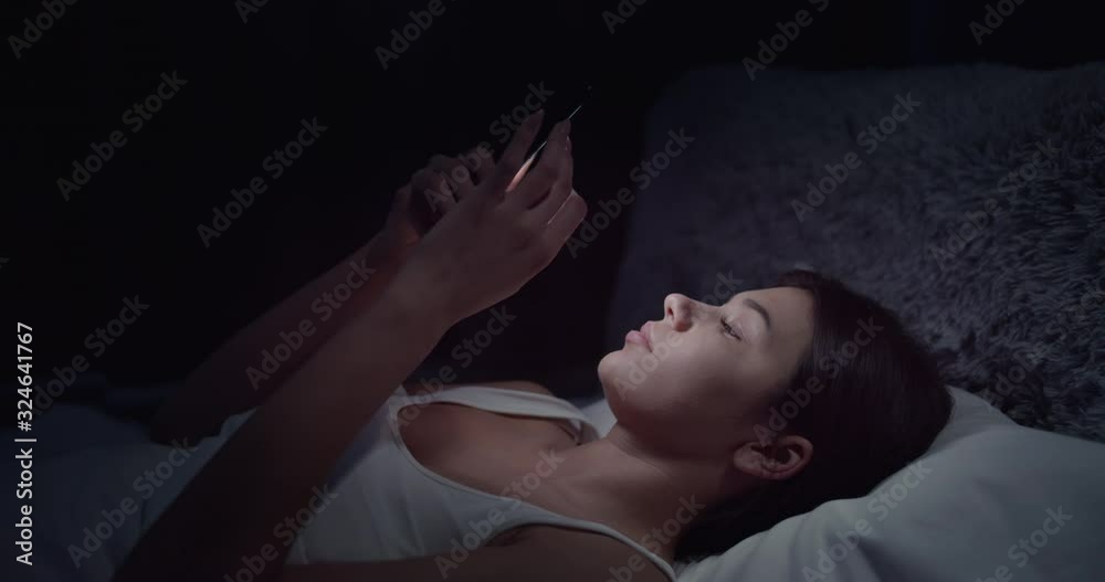 Pretty dark-haired woman lying in bed and scrolling smartphone in darkness, gadget addiction