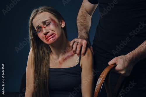Male put his hand on female shoulder, showing belt. Victim with bruises on face sitting nearby, crying. Blue background. Domestic violence. Close-up.