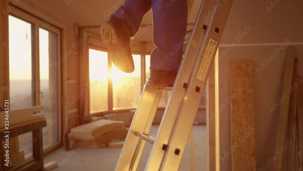 SLOW MOTION, CLOSE UP, LENS FLARE, DOF: Builder in blue overalls climbs down a aluminium ladder at sunset. Unrecognizable construction site worker climbs down an extendable ladder on a sunny evening.